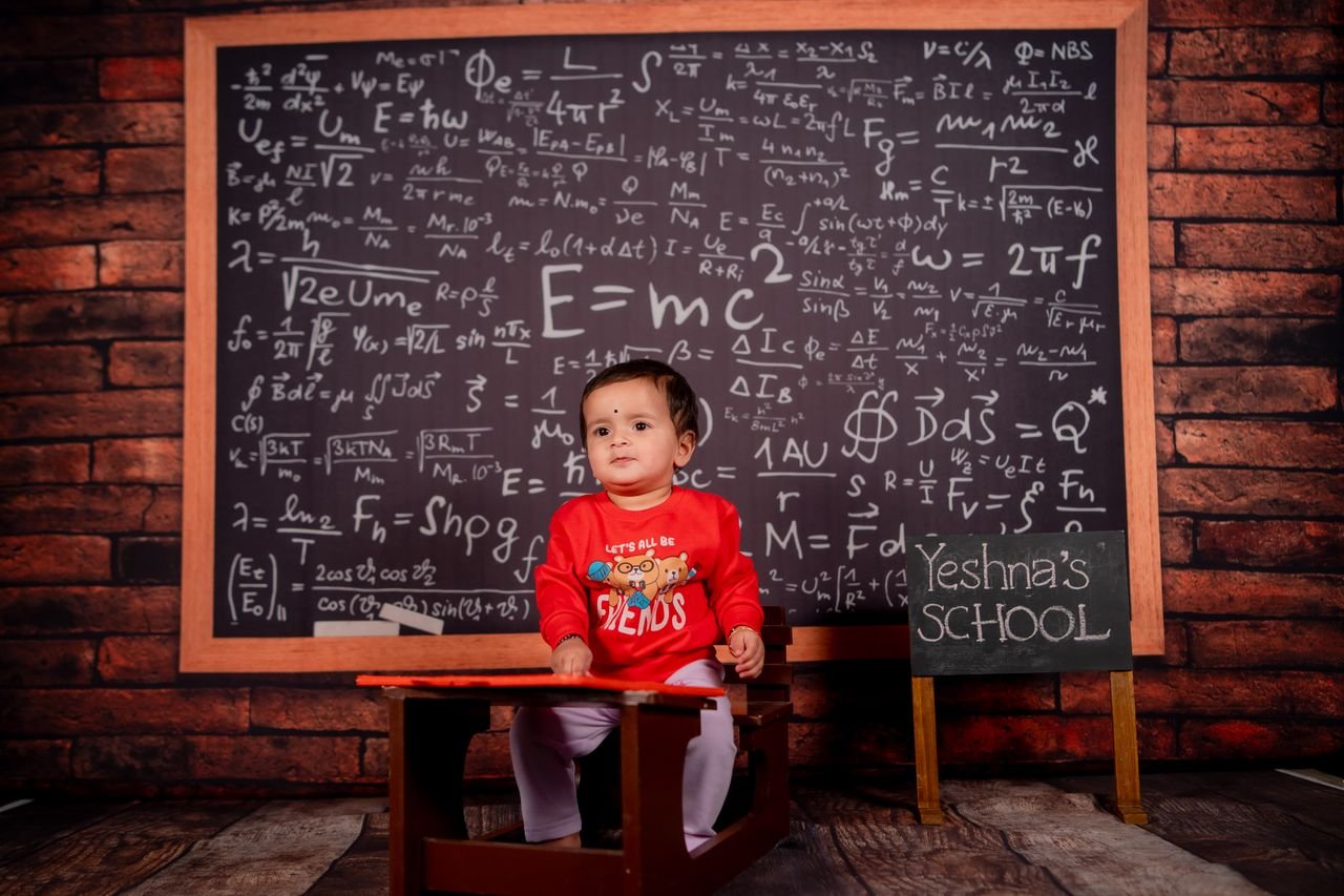 A baby in a classroom setting with a math-filled chalkboard background.