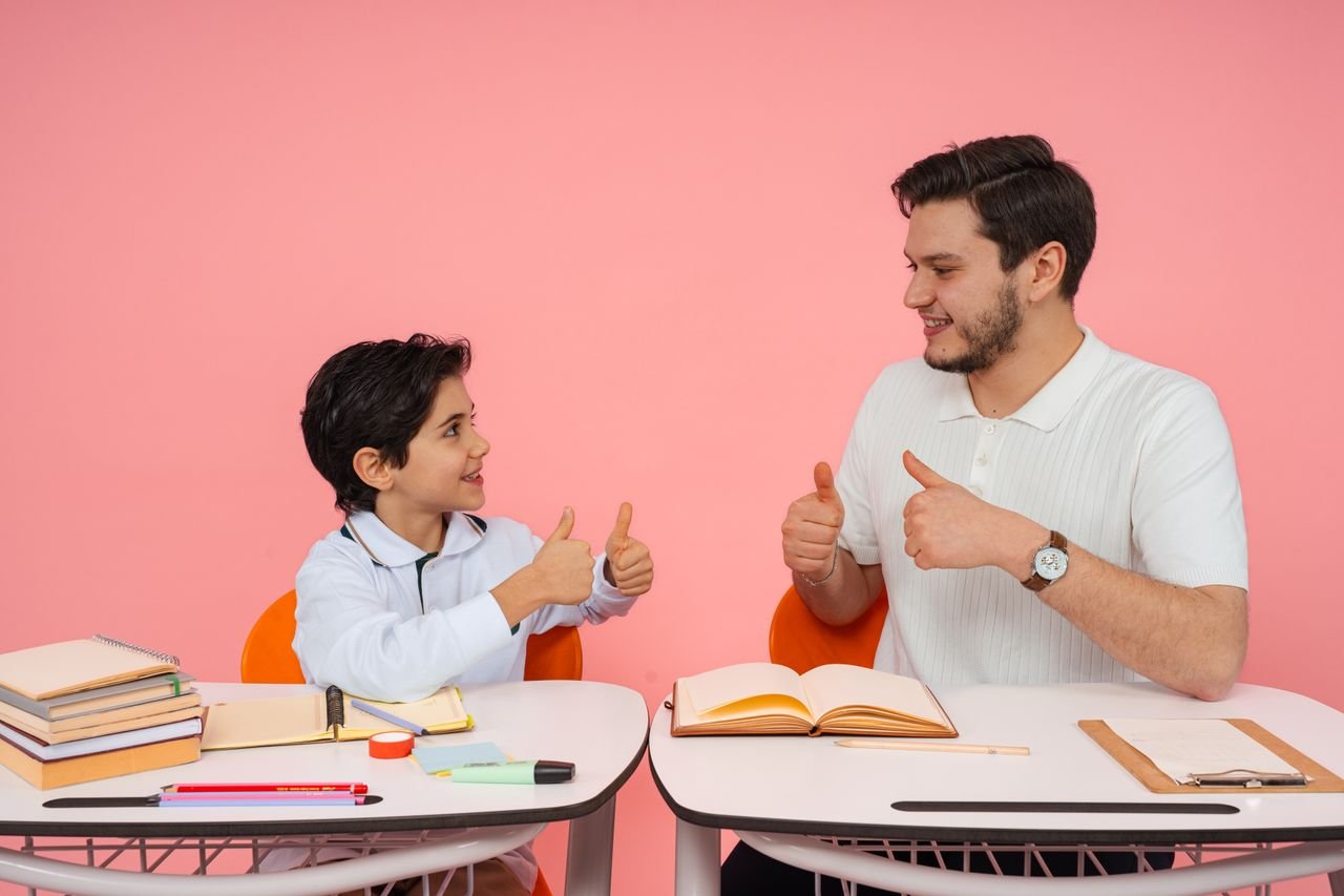 Teacher and student exchanging thumbs up at desk in a learning environment.