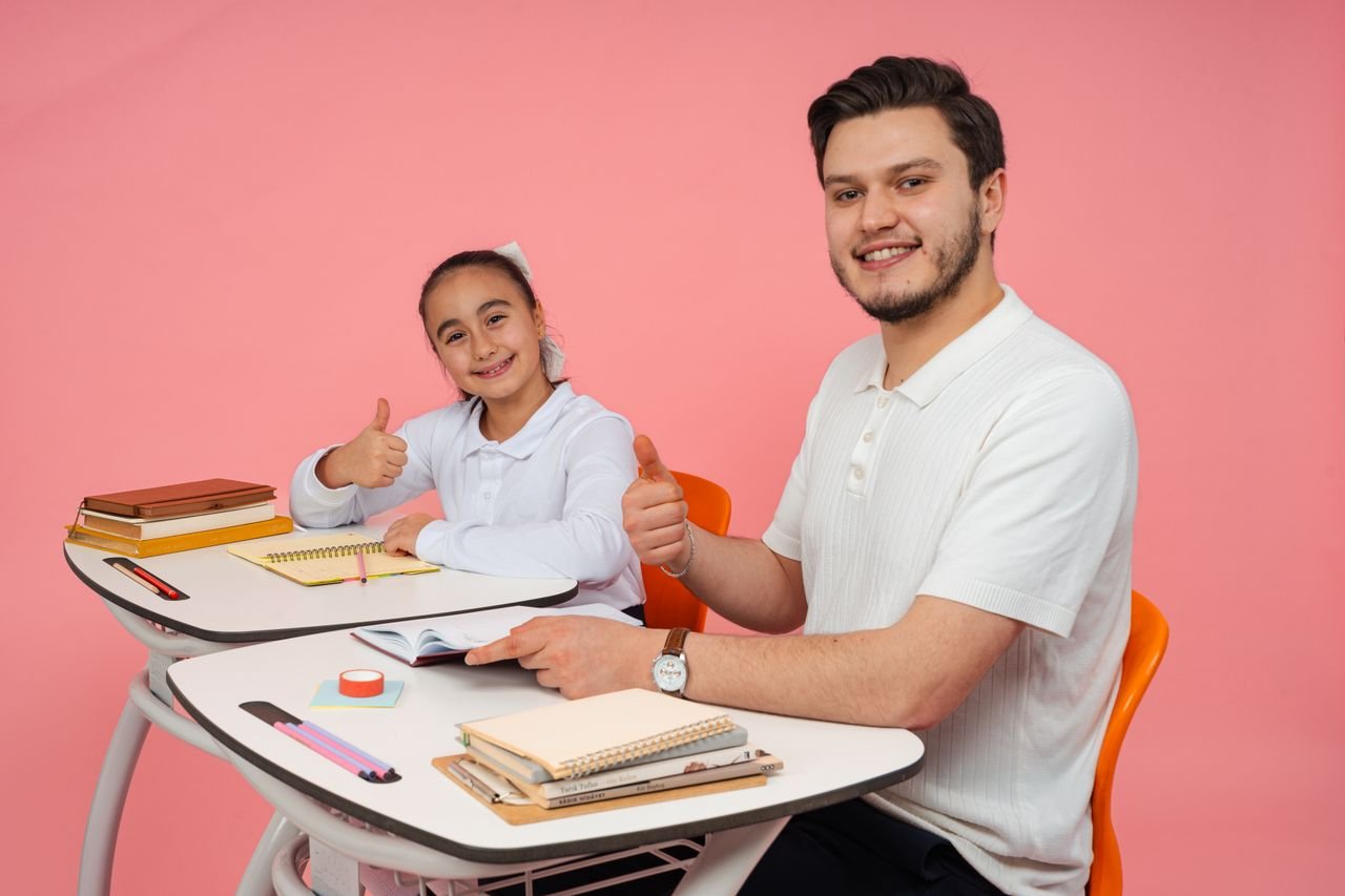 Smiling teacher and student at a desk, thumbs up, promoting education and positivity.