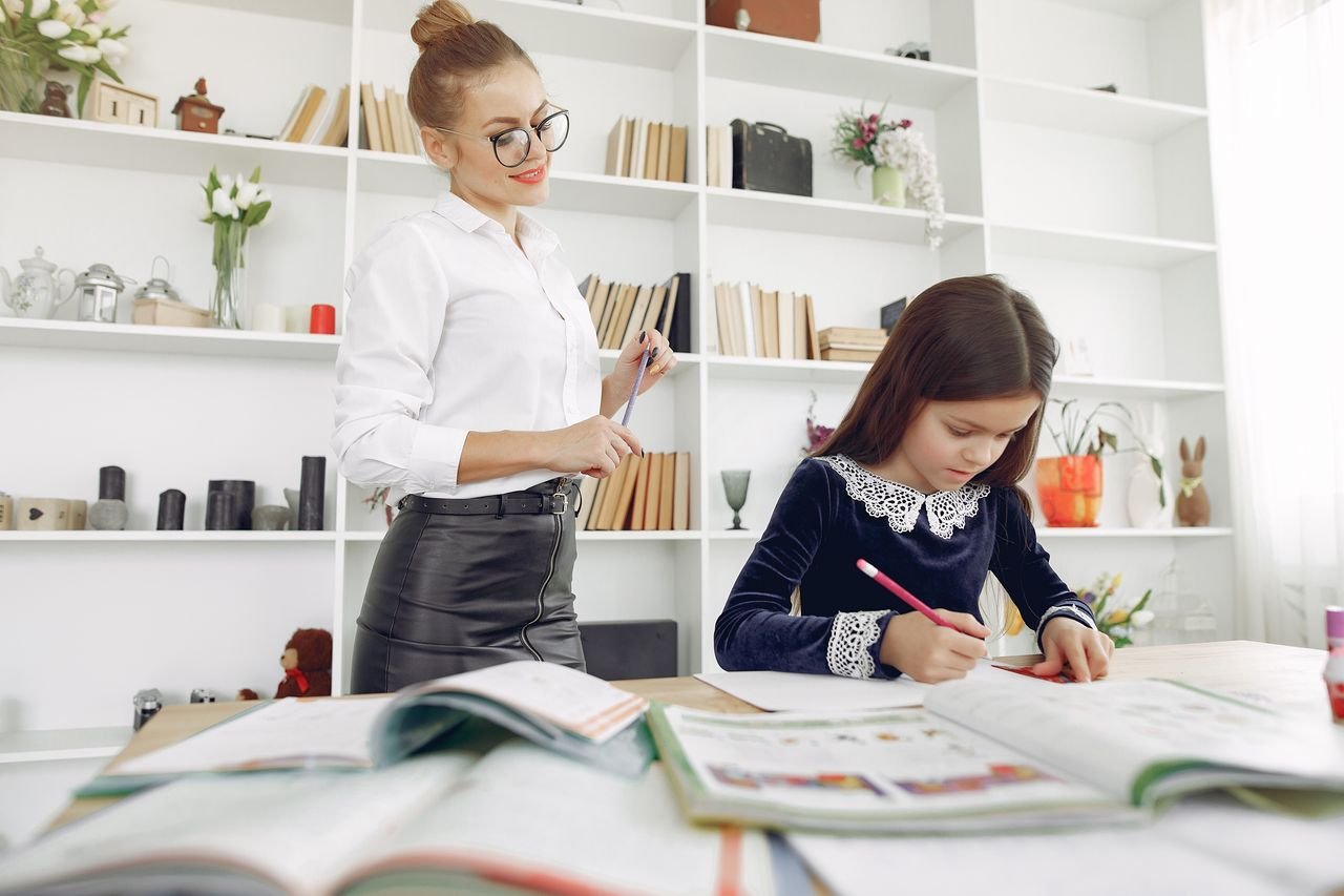 From below of focused girl sitting at table and doing homework tasks with content tutor in eyeglasses in creative place