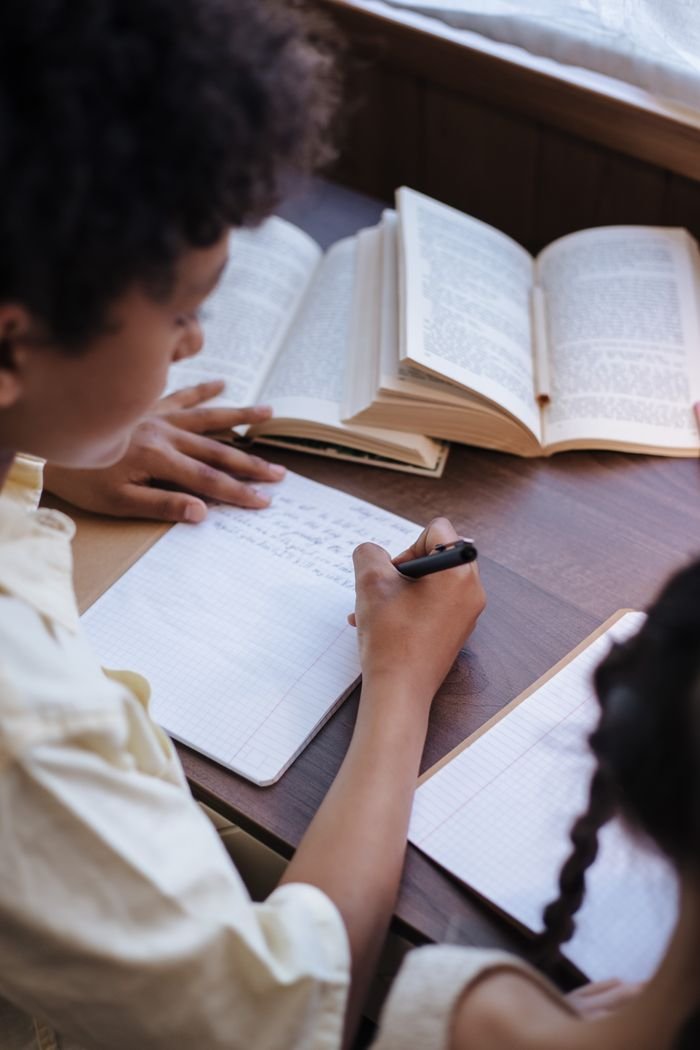 Two children writing in notebooks and reading books indoors, focused on learning together.